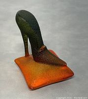 Side angle showing textured dark green to reddish-brown high heel shoe resting on orange gradient glass pillow base.