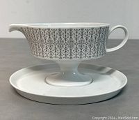 Side angle of the porcelain sauce boat on its matching saucer, clearly showing the black intricate geometric and floral design on a white background.
