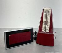 Two items: vintage red mechanical metronome and digital desktop alarm clock shown side by side on table.