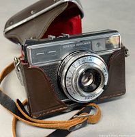 Camera in original brown leather carrying case with red fabric interior and strap, angled showing the front lens and markings.