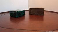 Two polished mineral trinket/jewel boxes on wood surface, one green malachite square box and one rectangular Capetown marble box.
