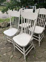Four white painted wooden dining chairs with spindle backs, decorative pressed top back panels, and acorn finials on top posts. One chair differs slightly in acorn finial design.