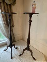Pair of Georgian-style tall wooden table stands with round tops and tripod bases, shown on a white cloth near windows with curtains.