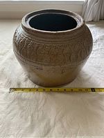 Front view of brown antique Chinese olive jar showing carved pattern and round opening with measuring tape indicating width approx 10 inches.