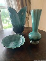 Three ceramic pieces on a wooden table near window light, showing leaf-shaped vase, tall teal vase, and shallow green dish.