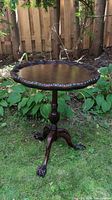 Full view of the round wooden occasional table with carved claw feet, pedestal, and scalloped edge outside on grass.