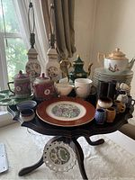 Assorted Chinese porcelain and ceramic tableware arranged on a small wooden table by a window. Items include teapots, mugs, plates, small bowls, condiment jars, and serving dish.