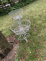 Full view of the two-tier round pot holder made of white painted metal with filigree rod design and three curled legs, placed outside on grass near a tree.