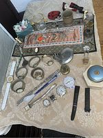 Overview of watches, compacts, embroidered mat and assorted small decorative items on a beige patterned fabric.