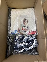 Box open showing folded clothes including white graphic tee and blue/white shirt underneath