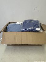 Box containing folded clothing items in blue and grey tones, some new and sealed in plastic.