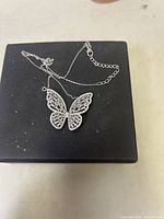 Detailed view of butterfly pendant necklace laid flat on a black box, showing the pendant and fine chain with clasp and extender chain visible.