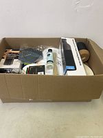 Overview photo of cardboard box containing various household and kitchen items including a bottle of liquid, small boxes, kitchen tools and a cork handled mug.