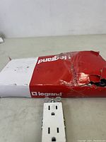 Two images showing a white Legrand duplex electrical outlet socket in front of a red and white Legrand branded box. The box is damaged.