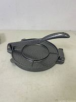 Top and angled side view of gray metal tortilla maker press with handle lifted to show the pressing surface and hinge.