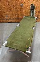 Two folding aluminum military cots with green fabric laid out on concrete floor against a plywood wall.