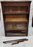 Front view of four stacked oak barrister book cases with glass panels, showing signs of wear and missing glass panel for one section. Wooden parts on floor in front of cases.