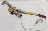 Overview of the steel ratchet rope puller with safety catch hooks, cable, and red handle grip laid out on a white cloth background.