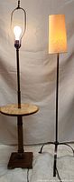 Full view of both floor lamps showing the MCM wooden lamp with built-in marble side table and exposed bulb, alongside a modern metal floor lamp with fabric shade and tripod base.