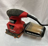Front angled view of red and black SKIL 7292 palm sander with attached micro-filtration dust canister showing the sanding base and dust filter.