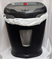 Front view of black and silver Staples high speed shredder with trash bag fitted inside, showing transparent window and wear marks at the base