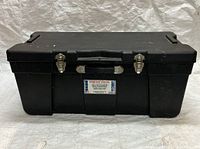 Front view close-up of black plastic storage trunk with two metal latches and carrying handle. Label on front center shows Terence Thompson Inc. branding and dimensions.