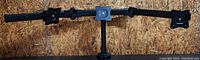 Full view of black triple monitor stand designed to hold three monitors mounting plates visible on two ends and one in the center, mounted on horizontal arms attached to central vertical pole with clamp base.