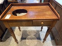 Front view of antique wooden washstand showing drawer, circular cutout on top, and turned legs.