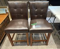 Two brown upholstered bar chairs with button tufted backs, wooden legs and footrests placed side by side on carpeted floor.