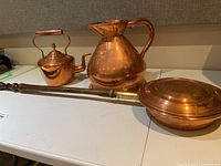 Photo of three copper items on white surface: 2 gallon jug, small teapot with lid, bed warmer with wooden handle
