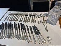 Wide view of the incomplete flatware set including forks, knives, spoons, with glass decanter and metal vase at right end.