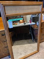 Full view of rectangular wall mirror with wooden frame and woven accent panel on top, standing against furniture.