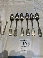 Six silver teaspoons and one pair of metal sugar cube tongs laid on white fabric.