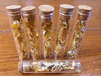 Photo of six clear glass vials containing gold leaf, sealed with cork stoppers, arranged standing on a wooden surface with one vial lying down.
