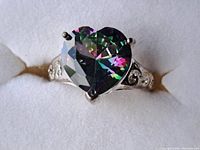 Close-up image of a sterling silver ring with a large heart-shaped mystic topaz gemstone showing multicolor iridescence and filigree details on band sides.