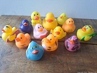 Photo displaying 13 assorted rubber ducks in various colors and styles on a wooden surface with a plain background.