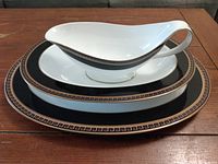 Three serving dishes stacked: a gravy boat on top of a vegetable bowl on top of a serving platter, featuring black and gold Greek key design on white fine bone china