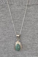 Sterling silver necklace displayed against a gray knit fabric background, showing the full length of the chain and prominently the green aventurine pendant in a silver setting.