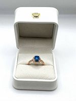 Blue opal ring displayed in white leatherette box with gold crown emblem.