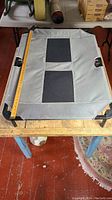 Top view of the elevated dog bed showing the gray fabric with two black mesh strips and metal frame with handles on the sides. Measuring tape shows width approximately 27 inches.