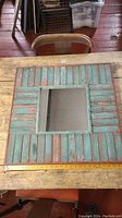 Full view of square wooden framed mirror with distressed paint and a mirror center on a work table.