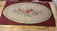 Full view of the rectangular hooked rug showing the burgundy border surrounding a beige field with a light green oval and central floral basket design.