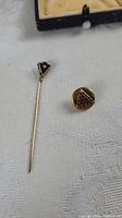 Two Masonic pins on white fabric, one stick pin with black triangular top and one smaller pin with triangular face and red dots.