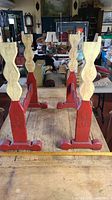 View of the entire wooden stand showing four cream painted hourglass-shaped posts on a red base with shoe feet, positioned upside down on a wooden table.