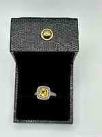 Ring with yellow sapphire center stone and white sapphire halo inside black velvet box with textured exterior and gold-tone button.