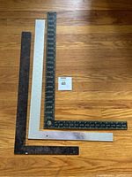 Three metal carpenter squares laid out flat forming right angles on wooden floor, showing overall shape and size.