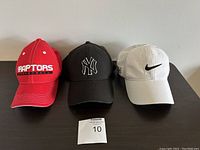Front view of three youth baseball caps in red, black, and white, showing logos of Toronto Raptors, New York Yankees, and Nike.