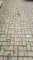 Long wooden pole with pruning blade, rope, and wooden handle grip on paving stones.