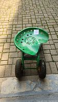 Front and overhead view of green metal perforated tractor-style garden seat with black plastic tray underneath and two visible pneumatic tires with yellow rims.