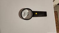 Black handheld magnifying glass with round lens and light button on handle, placed on white surface.